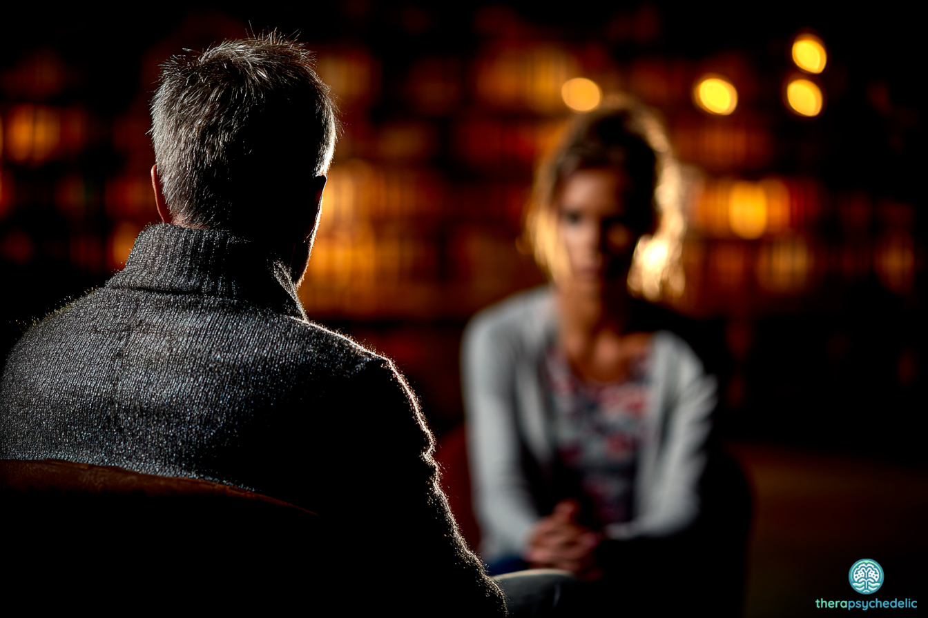 Un thérapeute, vu de dos, est assis et écoute attentivement une patiente assise en face de lui, de face et légèrement floue. La scène se déroule dans un environnement tamisé avec des lumières chaudes en arrière-plan, suggérant une atmosphère d'écoute et de discussion sereine. L'image évoque l'importance de l'accompagnement et du cadre thérapeutique.